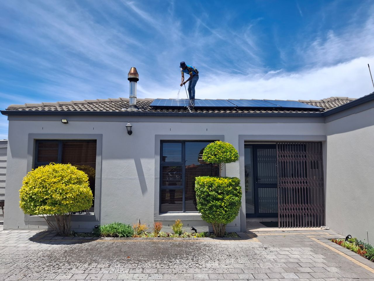 About A person maintains solar panels on a house roof under a clear blue sky.